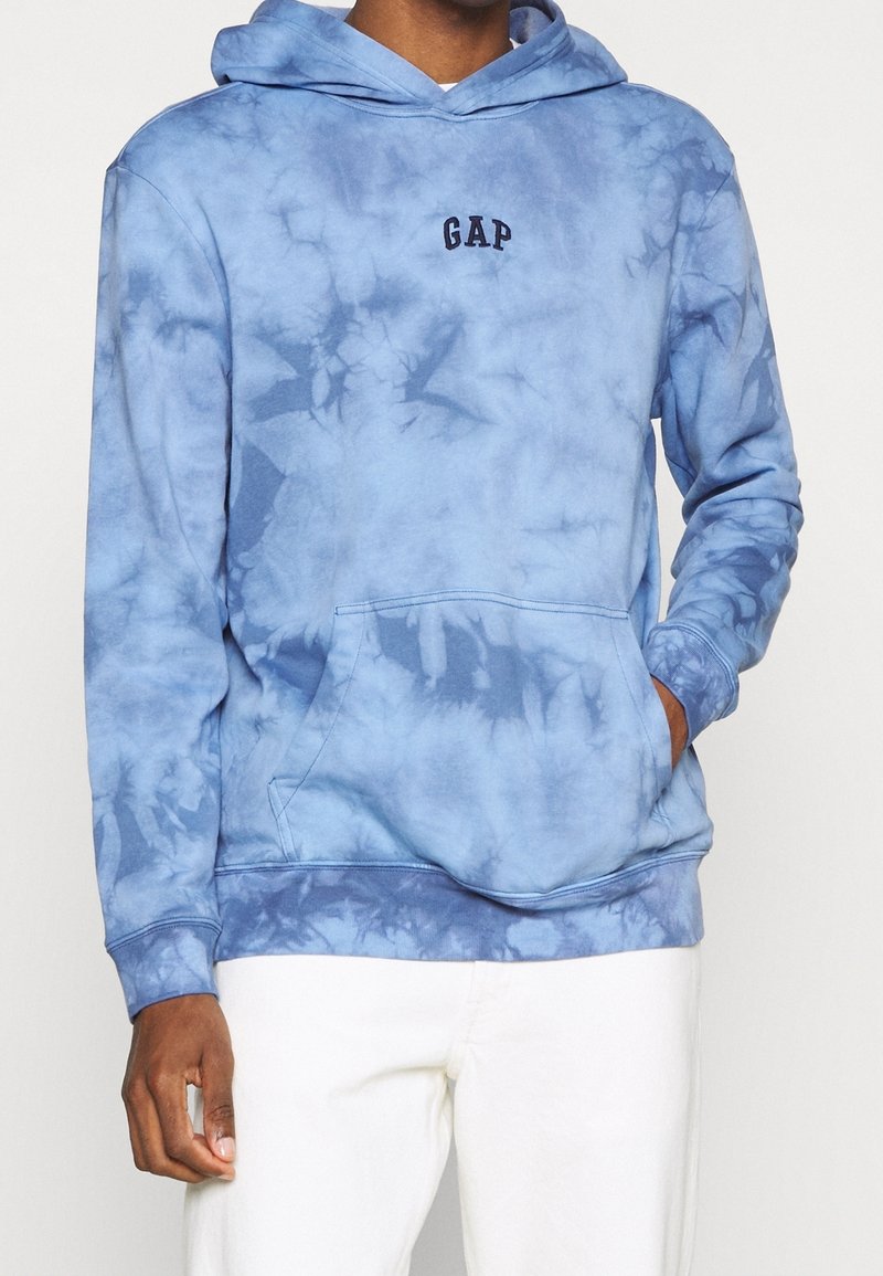 Homme portant un sweat à capuche bleu tie-dye GAP avec poche avant et pantalon blanc, debout devant un fond uni.