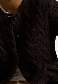 Mörkbrun kabelstickad cardigan med ett texturerat mönster, med svarta knappar och en liten broderad logotyp på bröstet.