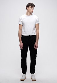 Biała koszulka z krótkim rękawem; czarne spodnie ze ściągaczem; białe trampki z beżowymi akcentami; model stoi prosto na jednolitym tle.