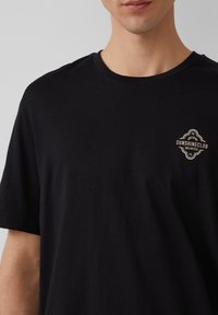 Schwarzes Baumwoll-T-Shirt mit Rundhalsausschnitt und kurzen Ärmeln. Verfügt über ein kleines cremefarbenes Logo im linken Brustbereich. Glatte Textur.