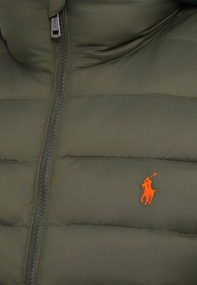 Maslinasto zelena pufasta prsluk s visokim ovratnikom, s prednjim zatvaračem i malim narančastim izvezenim logotipom konjanika s lijeve strane.