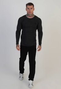 Black long-sleeve shirt with a textured pattern, paired with black pants and white sneakers. Model stands against a plain background.