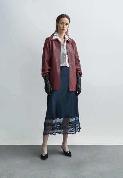 Γυναίκα που φοράει δερμάτινο μπουφάν σε βουργουνδί απόχρωση, ριγέ πουκάμισο, midi φούστα με δαντέλα σε ναυτικό μπλε, μαύρα γάντια και μαύρες γόβες, στέκεται σε γκρι δάπεδο.