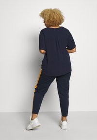 Navy top with short sleeves and back pleat, paired with navy pants featuring orange side stripes. Worn with white sneakers.