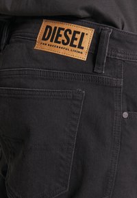 Close-up of black denim jeans back pocket with brown leather Diesel brand patch and belt loop detail.