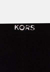 Schwarzer Stoff mit silbermäßig reflektierendem "KORS"-Text am Bund, wahrscheinlich Teil eines Kleidungsstücks wie Hosen oder Shorts.