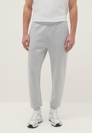 Uomo che indossa pantaloni della tuta grigio chiaro, scarpe da ginnastica bianche e calzini bianchi, in piedi davanti a uno sfondo chiaro e uniforme.