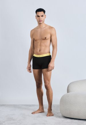 Jeune homme debout pieds nus sur un tapis, portant un caleçon noir avec une ceinture jaune Tommy Hilfiger, à côté d'un fauteuil blanc arrondi.