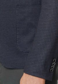 Blazer de color marino con un pequeño patrón de cuadrados, textura de tela y un bolsillo en el pecho. Acabado con cuatro botones negros en el puño.
