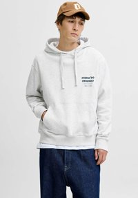 Sudadera gris claro con un bolsillo frontal y cordones, que presenta un texto gráfico en verde oscuro. Llevada con vaqueros de mezclilla azul oscuro. Gorra marrón.