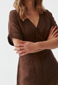 Braunes Leinenkleid mit V-Ausschnitt, kurzen aufgerollten Ärmeln und tailliertem Schnitt. Verfügt über eine dezente Textur und klare Linien.