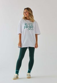 Biały oversize'owy t-shirt z zielonym napisem, w połączeniu z zielonymi legginsami i białymi trampkami. Skarpety mają akcenty w kolorze turkusowym i białym w paski.