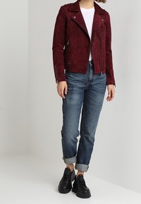 Bordeaux suède motorjack met zilveren ritsen, gedragen over een wit T-shirt. Gecombineerd met donkerblauwe spijkerbroeken met opgerolde mouwen en zwarte chunky sneakers.