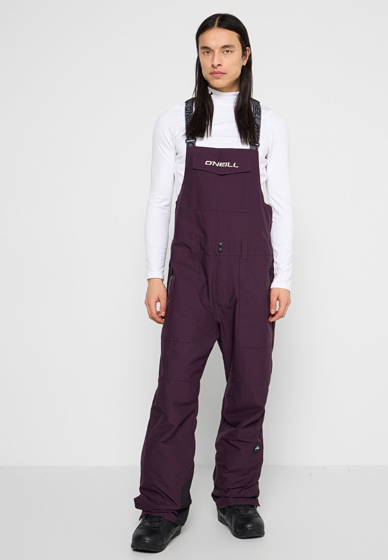 Fioletowe wodoodporne ogrodniczki śnieżne z regulowanymi ramiączkami, przednią kieszenią i wzmocnionymi mankietami, zestawione z białą bluzą z długim rękawem i czarnymi butami.