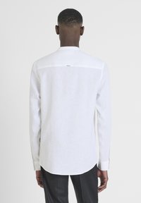 Camicia bianca a maniche lunghe in lino, con colletto alto, dettagli con bottoni sui polsini e una pence posteriore con un gancio per appendere.