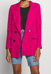 Fuchsiové sako s výrazným střihem, klopy s výřezem, dvouřadé knoflíky, boční kapsy, nošené přes černý top a džínové kraťasy.