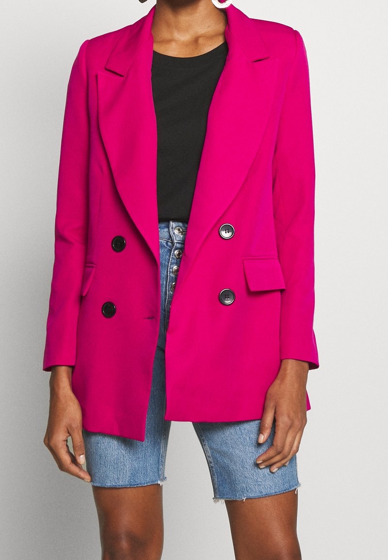 Fuchsiové sako s výrazným střihem, klopy s výřezem, dvouřadé knoflíky, boční kapsy, nošené přes černý top a džínové kraťasy.