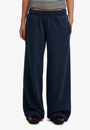 Osoba nosząca luźne granatowe spodnie dresowe z elastyczną talią, krótki pasiasty top i ciemne buty na białym tle.