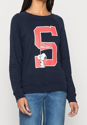 Persona vistiendo un suéter azul marino con una gran letra roja "S" y Snoopy sosteniendo una bandera roja que dice "¡RAH RAH!" en el frente.