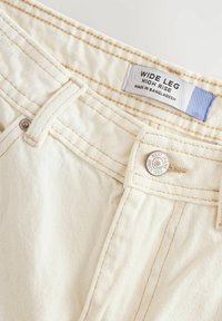 Bredde ben, høj talje, cremefarvede denimjeans med tekstureret stof, guldtråde og en sølvknap med brandets påskrift.