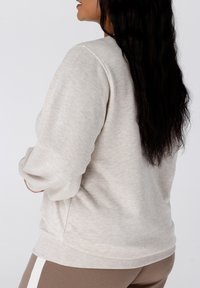 Femme aux longs cheveux noirs portant un sweat-shirt beige clair et un pantalon brun avec une rayure blanche, de profil devant un fond uni.