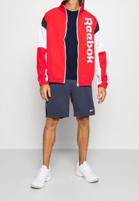 Röd och vit zip-up jacka med en stor logotyp, matchad med marinblå shorts. Den kompletta outfiten inkluderar vita sneakers. Minimalistisk design.