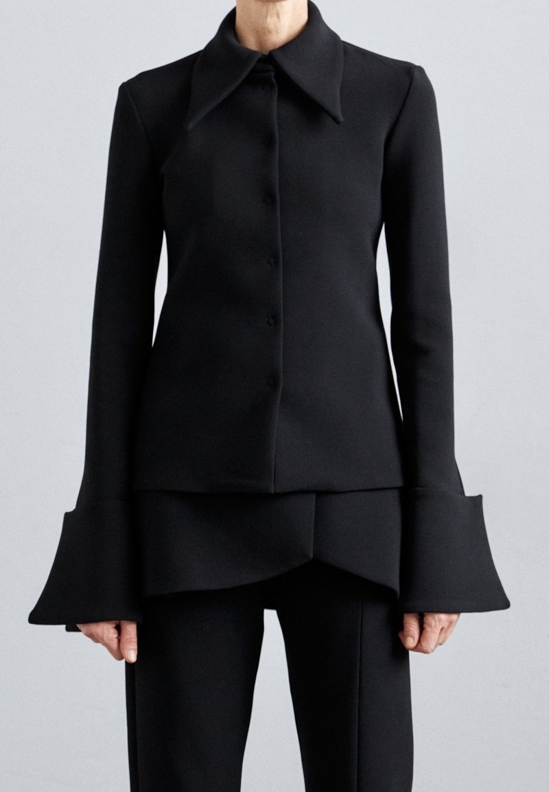 Femme portant une veste noire cintrée avec des poignets évasés et un pantalon assorti sur un fond clair uni.