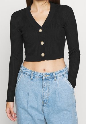 Žena v černém žebrovaném krátkém kardiganu s knoflíky a světle modrých džínách s vysokým pasem, s rukama v kapsách.