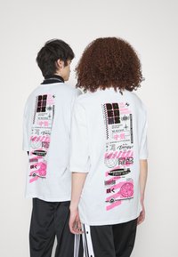 Bílý oversize tričko s grafickými potisky v černé a růžové na zadní straně, obsahující různé texty, kruhové a květinové vzory a výrazné akcenty.