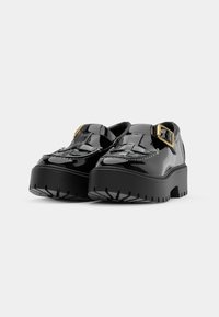 Sapatos de plataforma em couro sintético preto com sola espessa, apresentando uma tira decorativa e detalhe de fivela dourada de um lado.