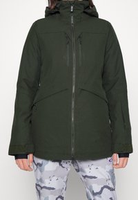 Giacca impermeabile verde scuro con un design aderente, due tasche sul petto e cappuccio regolabile. Indossata sopra pantaloni grigi con motivo camouflage.