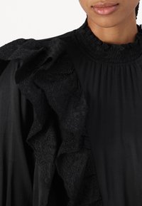 Blusa negra con detalles de mangas rizadas texturizadas, que presenta un bordado intrincado y un escote fruncido. Tejido suave, silueta elegante.