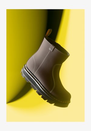 Bruine enkellaars met witte stiksels, gestructureerde bovenkant en dikke zwarte zool met robuust profiel. Geel label aan de achterkant.