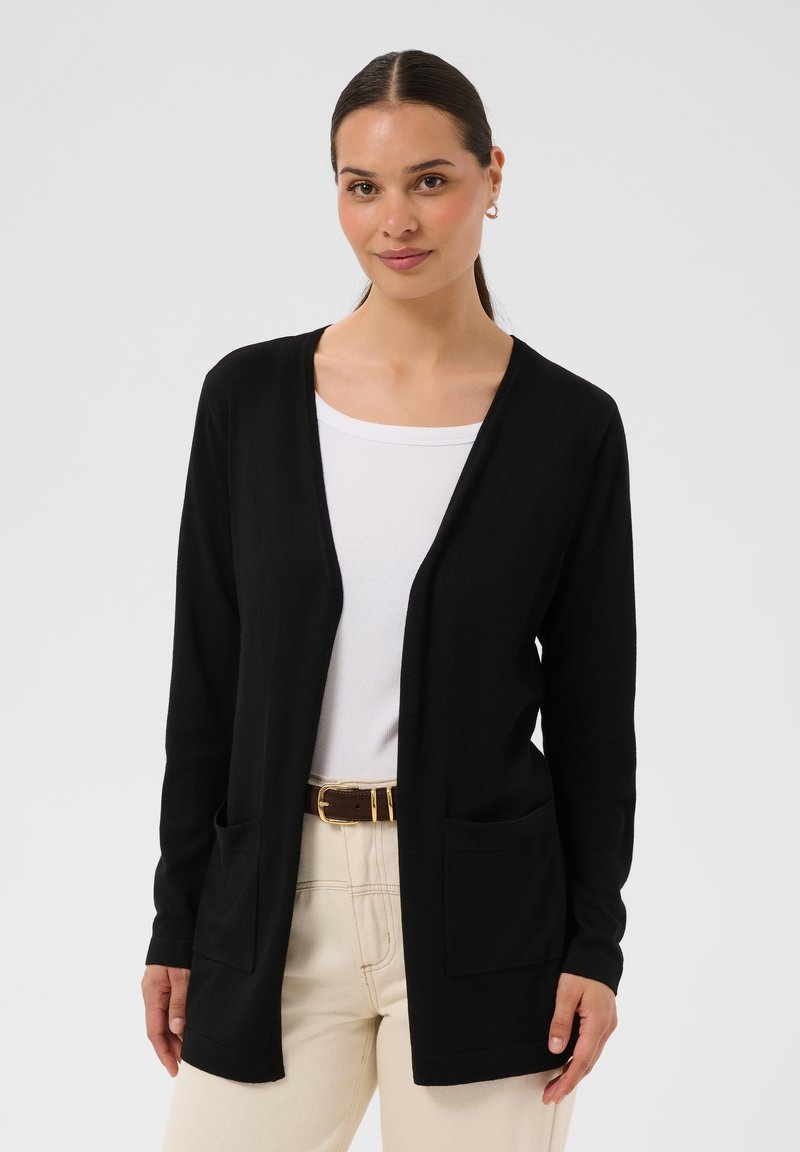 Femme portant un cardigan noir ouvert sur une chemise blanche, pantalon beige avec une ceinture marron, debout devant un fond clair uni.