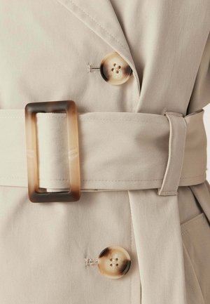 Trench beige con cintura abbinata chiusa da fibbia rettangolare in tartaruga e due grandi bottoni rotondi in tartaruga.