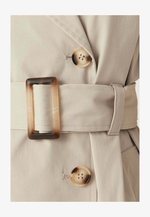Beige trenchcoat met bijpassende riem, vastgemaakt met een rechthoekige schildpadsluiting en twee grote ronde schildpadsknopen.