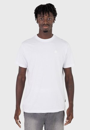 Ung mann med korte dreadlocks iført en ensfarget hvit kortermet t-skjorte og grå bukser, stående foran en lys grå bakgrunn.