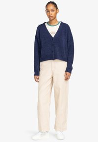 Marineblauwe cropped cardigan met een zachte textuur, featuring een V-hals, vijf zwarte knopen en verlaagde schouders, gedragen met crèmekleurige ruime broek.