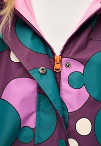 Zbliżenie na wielokolorową kurtkę z okrągłymi wzorami w kolorach morskim i fioletowym, pomarańczowym zamkiem błyskawicznym, zatrzaskami oraz białym golfem pod spodem.