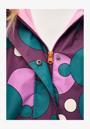 Zbliżenie na wielokolorową kurtkę z okrągłymi wzorami w kolorach morskim i fioletowym, pomarańczowym zamkiem błyskawicznym, zatrzaskami oraz białym golfem pod spodem.