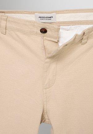Beżowe spodnie z częściowo rozpiętym rozporkiem na guziki, szlufkami na pasek i metką "JACK & JONES PANTS STUDIO" wewnątrz paska talerza.