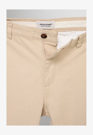 Beżowe spodnie z częściowo rozpiętym rozporkiem na guziki, szlufkami na pasek i metką "JACK & JONES PANTS STUDIO" wewnątrz paska talerza.