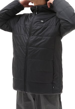 Personne ajustant le col d'une veste matelassée noire Vans zippée, portée avec un pantalon foncé, cadrée au niveau du menton et de la taille.