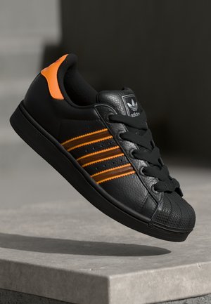 Czarny sneaker Adidas z pomarańczowymi paskami i detalem na pięcie, zasznurowany, przedstawiony nad szarą betonową krawędzią na ciemnym tle.