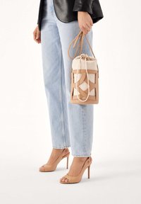 Beżowa torba sznurkowa z tkaniny z akcentami ze skóry, z geometricznym wzorem wycięć. Noszona z jasnoniebieskimi jeansami o prostych nogawkach i beżowymi szpilkami.