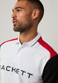 Polo de manga corta en blanco con detalles en rojo y negro, que presenta un cuello, una botonera y un gran logo negro de "HACKETT" en la parte frontal.