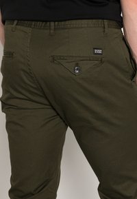 Pantalones de color verde oliva hechos de algodón, con un bolsillo trasero con botón y un diseño triangular. Material y construcción simples y duraderos.