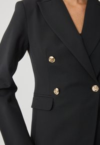 Μαύρο προσαρμοσμένο σακάκι με σχιστές απολήξεις, διπλό κούμπωμα και χρυσά, υφασμάτινα κουμπιά. Περιλαμβάνει λεπτομέρεια πλάγιας τσέπης.
