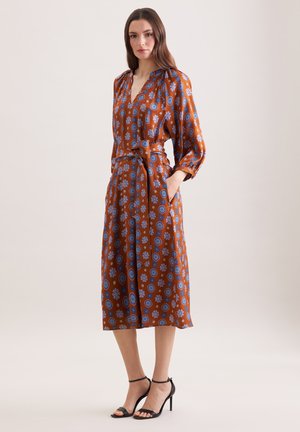 Donna che indossa un vestito midi color ruggine con motivi floreali blu, maniche lunghe, vita annodata e tacchi neri con cinturino alla caviglia, in piedi con una mano in tasca.