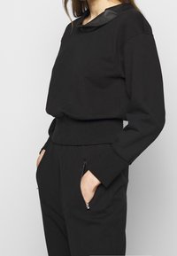 Černý top s dlouhým rukávem, se saténovým límečkem, žebrovaným pasem a kapsami. Hladká textura, volný střih a ploché švy.